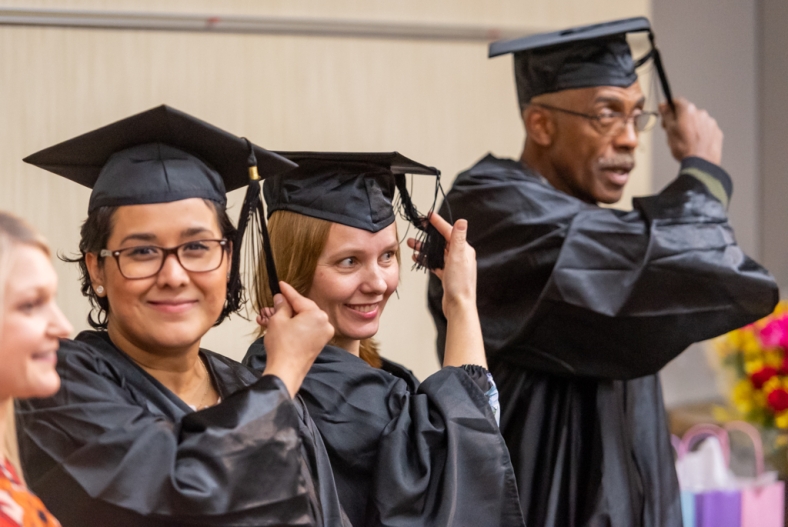 Three adults dressed in black graduation caps and gowns prepare to move their tassles. A Latina woman gazes directly into camera, standing next to a White woman and an older Black man.