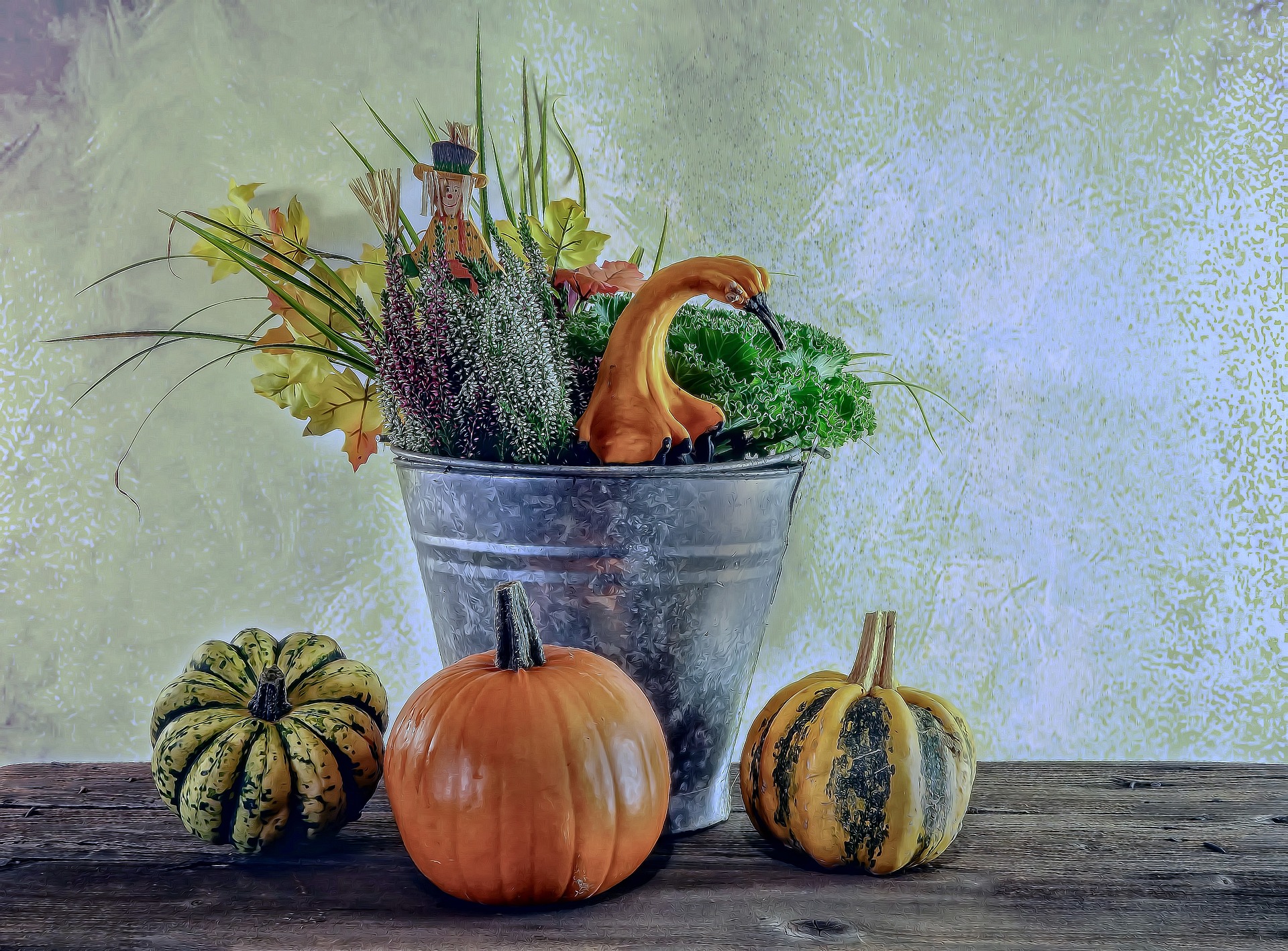 A metal watering pail holds fall greenery and herbs, and three small gourds sit on the wooden table at its base.