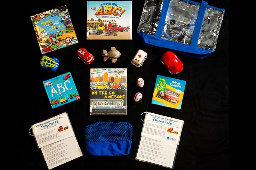 The contents of the On the Go Literacy Activity Kit displayed against black background, including toy vehicles, books, and tip sheets.